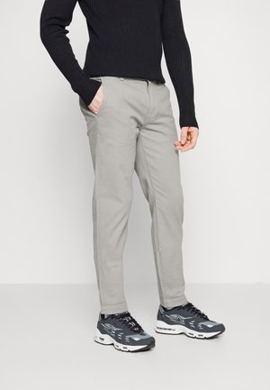 Person trägt hellgraue Hose, schwarzen gerippten Pullover und schwarz-weiße Sportschuhe und steht mit einer Hand in der Tasche.