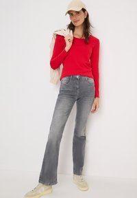 Röd långärmad topp, grå utställda jeans och beige sneakers. Modellen bär en beige keps och håller en ljusfärgad jacka över axeln.