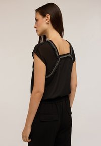 Zwarte top met korte mouwen en een vierkante achterkant, versierd met metallic trim langs de halslijn en de schoudernaden. Glad stoffen textuur.