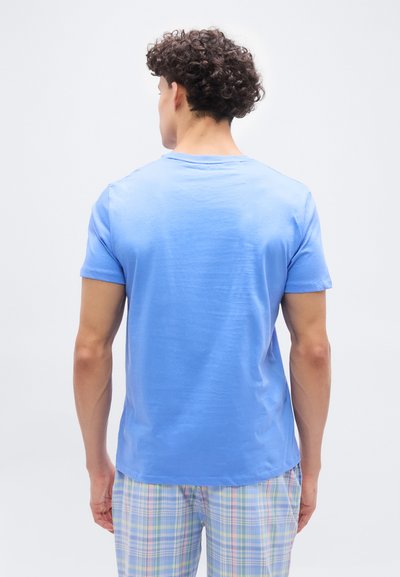 Άνδρας με σγουρά μαλλιά, που φοράει ανοιχτό μπλε κοντομάνικο μπλουζάκι και παντελόνι με παστέλ καρό σχέδιο, φαίνεται από πίσω μπροστά σε απλό φόντο.