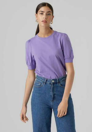 T-shirt court violet à manches courtes avec épaules bouffantes, en coton doux. Porté avec un jean bleu taille haute, fermetures à boutons visibles à l'avant.