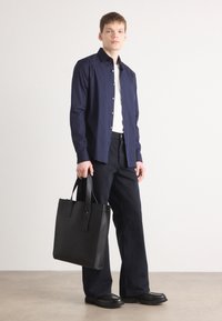 Cămașă albastră cu nasturi, tricou alb, pantaloni închiși, geantă de piele neagră cu mânere plate și încălțăminte neagră, pe un fundal neutru.