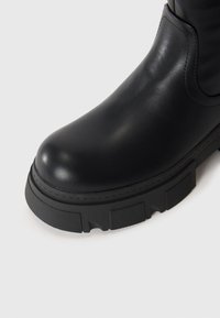 Bottes en cuir noir avec bout arrondi et semelle en caoutchouc texturé épais sur fond blanc.