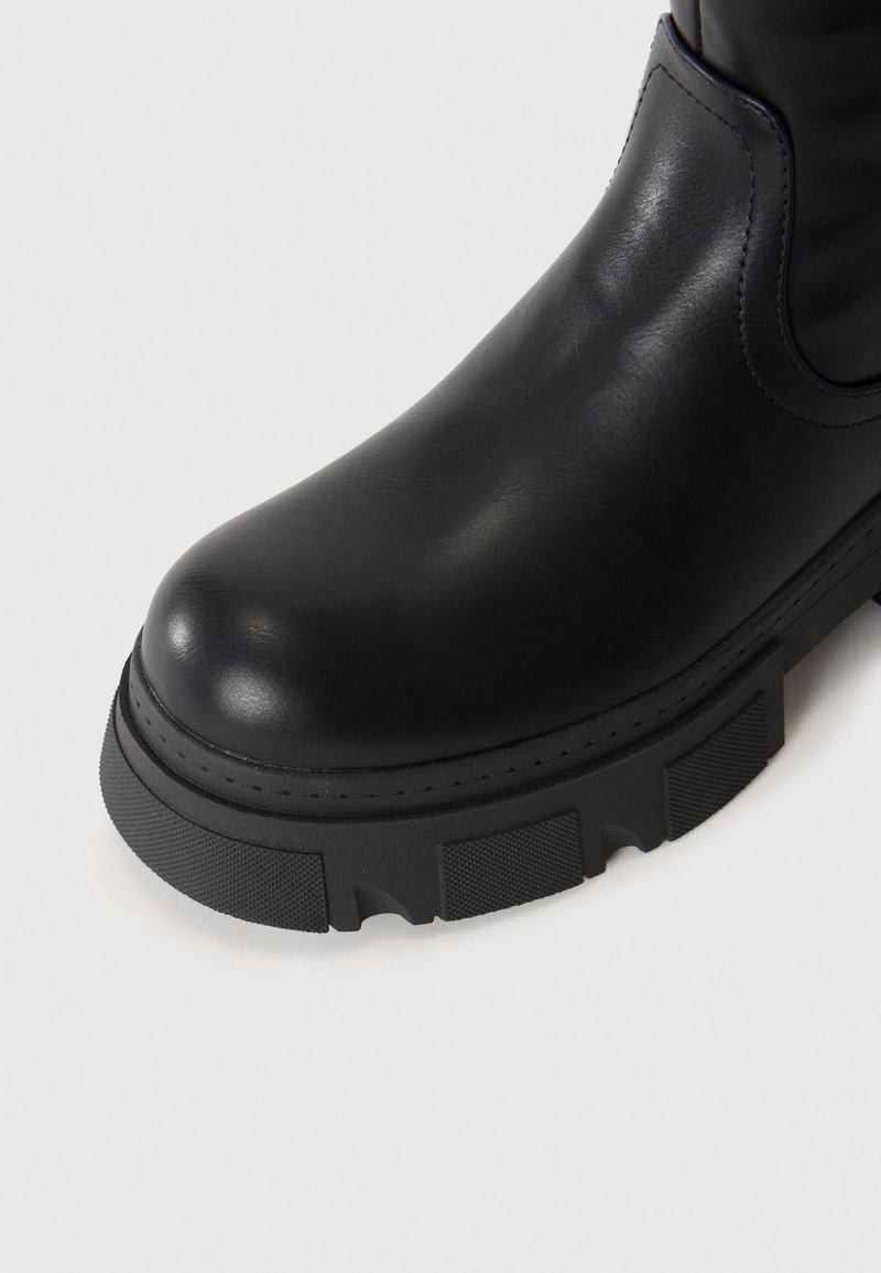 Zwarte leren laars met ronde neus en dikke, getextureerde rubberen zool op een witte achtergrond.