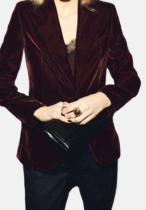 Donna che indossa un blazer in velluto rosso scuro sopra una canotta con bordo in pizzo, che tiene in mano una pochette nera con anelli alle dita.