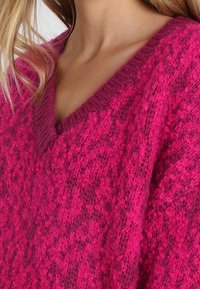 V-Ausschnitt-Pullover in leuchtendem Pink aus strukturierter Strickware mit lockeren, flauschigen Garnen. Der Stoff hat ein weiches, erhöhtes Aussehen mit unterschiedlichen Mustern.