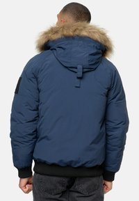 Blå polstret jakke med en faux fur-fórret hætte, ribbede manchetter og sort patchdetalje på ærmet. Har en afslappet pasform for varme.
