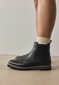 Mustad nahast brogue Chelsea saapad, kantud kreemja soonikkoega sokkidega, kujutatud heledal põrandal neutraalse tausta ees.