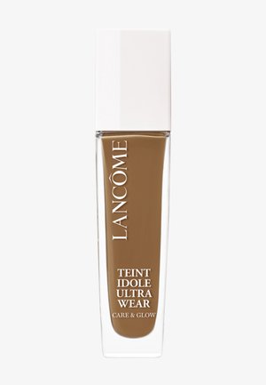Lancôme Teint Idole Ultra Wear foundation i mørkebrun. Klar glassflaske med et hvitt lokk, har elegant merkevarebygging og et enkelt design.