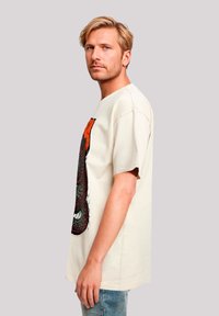 Beige T-Shirt mit kurzen Ärmeln aus Baumwolle, mit einem großen Grafikdesign in Rot und Schwarz, lockerer Schnitt und rundem Halsausschnitt.