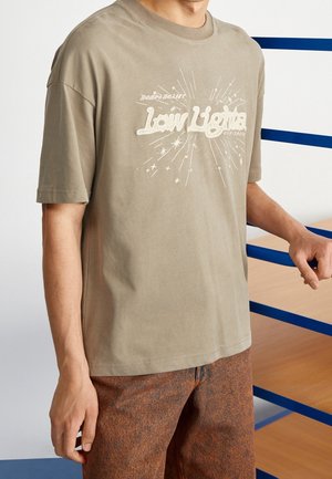 Man wearing a beige t-shirt with "Low Lights" text and starburst graphic, paired with brown textured pants, standing by a blue railing.