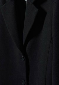 Manteau noir sur mesure avec larges revers et deux boutons visibles à la fermeture devant.