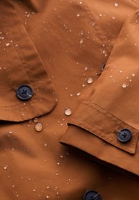 Brązowy wodoodporny materiał kurtki z kroplami, z czarnymi guzikami i detalami tekstury. Szwy widoczne na krawędziach i mankietach.