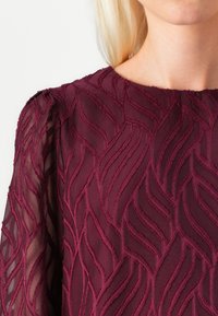 Burgundy dress with textured wavy pattern, featuring sheer sleeves and a crew neckline, highlighting intricate stitching details.