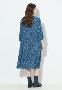 Sinivihreä kukkamekko, jossa on kolmannen neljänneksen hihat, pleatattu muotoilu ja polvipituinen helma, yhdistetty mustiin nilkkabootseihin. Silkkinen kangas.