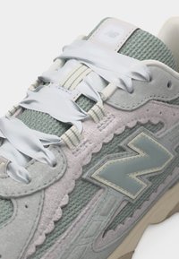 Κοντινό πλάνο ενός γκρι και ανοιχτοπράσινου αθλητικού παπουτσιού New Balance με λευκά φαρδιά κορδόνια, διχτυωτό επάνω μέρος, σουέτ επενδύσεις και μεγάλο λογότυπο "N" στη πλευρά.