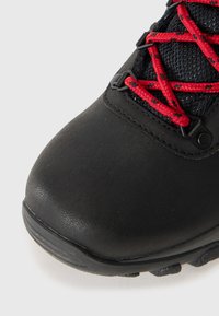 Chaussure de randonnée noire avec une tige en cuir texturé, semelle en caoutchouc épais et lacets en nylon rouge passés à travers une quincaillerie noire.