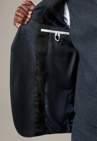 Interno di giacca blu scuro con tessuto strutturato, caratterizzato da una fodera in raso nero, cuciture bianche e una tasca con chiusura a bottone.