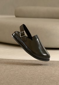 Μαύρο δερμάτινο παπούτσι slingback με ανοιχτή μύτη και ασημί αγκράφα. Λείο υλικό και στρογγυλεμένο σχήμα, με επίπεδη σόλα.