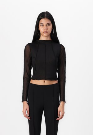 Top negro transparente de manga larga con cuello alto y diseño recortado que presenta sutiles detalles de costura. Combinado con pantalones negros ajustados.