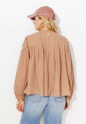 Blouse texturée couleur pêche avec des manches longues, ouverture en forme de trou de serrure au dos, et détails de couture décoratifs sur les épaules. Associée à un jean en denim bleu clair.