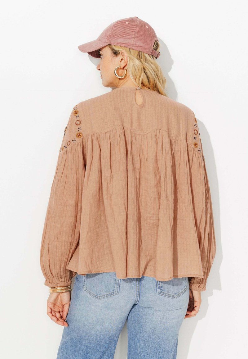 Blouse texturée couleur pêche avec des manches longues, ouverture en forme de trou de serrure au dos, et détails de couture décoratifs sur les épaules. Associée à un jean en denim bleu clair.