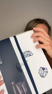 Hvit gaveeske med marineblå sløyfe; har preget bladmotiv. Esken inneholder tre mørkegrå tuber og en sprayflaske.