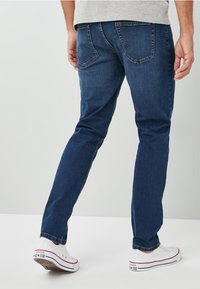 Jeans de denim azul oscuro con corte recto, que presentan dos bolsillos traseros y detalles de costura sutiles. Llevados con zapatillas blancas.