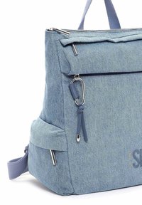 Ljusblå tygryggsäck med en texturerad denimfinish, framfick med dragkedja och silverfärgade metallaccenter. Har sidofickor och justerbara axelremmar.