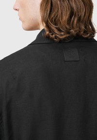Svart jacka med texturerad yta, med en slagkrage och en fyrkantig lädertagg som detaljerar "Alaïa" på baksidan.