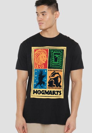 Mann trägt ein schwarzes T-Shirt mit bunten Hogwarts-Hausmaskottchen und dem Wort "Hogwarts" unter den vier Tiersymbolen.