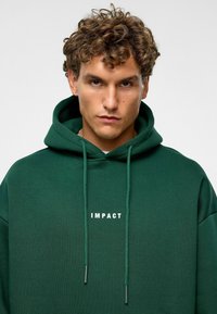 Pull à capuche vert en coton, avec une poche kangourou à l'avant, un cordon de serrage réglable et un logo textuel blanc "IMPACT" sur la poitrine.