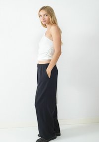 Blonde vrouw in een witte top met één schouder en losse zwarte broek staat zijwaarts met handen in de zakken tegen een witte muur.