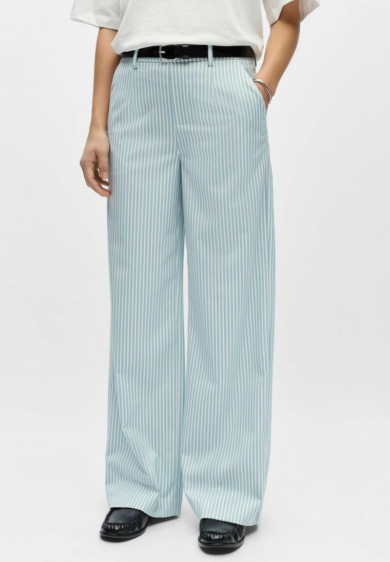 Helehallid ja valged triibulised laia säärega püksid, millel on sile tekstuur, must vöö ja küljetaskud, kantud mustade kingadega.