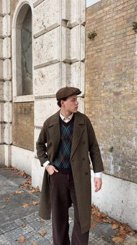 Abrigo marrón a cuadros, chaleco azul marino con diseño de rombos, camisa blanca, pantalones marrones de pana y gorra plana marrón, con una pared de piedra texturizada de fondo.