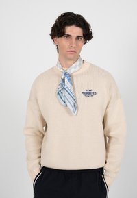 Pull en maille beige avec des poignets côtelés, arborant un logo "PROHIBITED" brodé en bleu. Associé à une écharpe bleue et blanche à motif autour du cou.