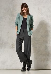 Ljus turkos stickad cardigan med knappar, bärs över en svart topp, i kombination med grå vida byxor och svarta ankelfutter. Texturerad bakgrund.