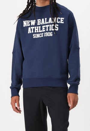 Mann trägt dunkelblauen Sweatshirt mit weißem Schriftzug "NEW BALANCE ATHLETICS SINCE 1906", kombiniert mit schwarzer Hose, steht vor weißem Hintergrund.