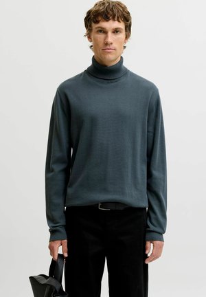 Jeune homme aux cheveux courts et bouclés, portant un pull à col roulé gris foncé et un pantalon noir, tenant un sac noir sur un fond uni.