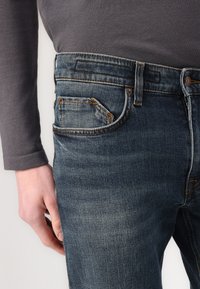 Sötétkék farmernadrág, kopott textúrával, egy kis elülső zsebbel és narancssárga varrásokkal. A derék alacsony szabású, övhurkokkal.