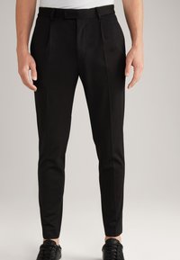 Schwarze maßgeschneiderte Hose mit schlankem Schnitt, ausgestattet mit Falten und Gürtelschlaufen. Getragen mit einem weißen Hemd und schwarzen Sneakers. Glatte Textur.