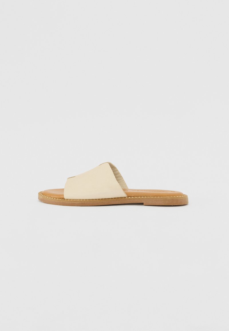 Beige slide sandal with a soft fabric upper, cross-over design, and a brown textured footbed, featuring a decorative stitch along the edge.