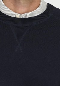 Suéter de punto azul marino con cuello redondo, con un detalle de punto cruz en el cuello y puños y dobladillo acanalados. Un cuello a rayas asoma por debajo.