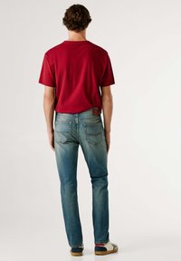 Rød t-skjorte med korte ermer, blå denimjeans med falmet finish, og fritidssko med blått og beige design, sett bakfra.