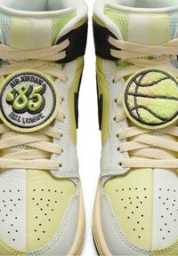 Paire de baskets montantes jaunes et blanches avec des lacets beige, chacune présentant des patchs brodés ronds : l'une avec le texte "Air Jordan '85", l'autre avec un ballon de basket.