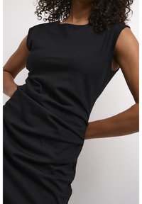 Femme aux cheveux bouclés portant une robe noire sans manches avec un tissu froncé, debout devant un fond clair uni.