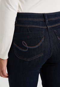 Jean en denim bleu foncé au design ajusté, avec des coutures orangées visibles et une poche arrondie accentuée à l'arrière.