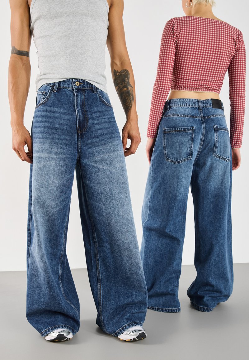 Weite blaue Denim-Jeans mit einem verwaschenen Muster, großen Taschen und einem nahtlosen Design. Getragen mit einem grauen Tanktop und einem rot-karierten Cropped-Top.