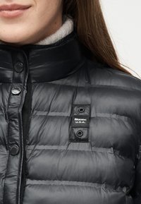 Gros plan d'une personne portant une doudoune noire matelassée avec des boutons-pression et une étiquette "Blauer U.S.A." sur la poitrine.