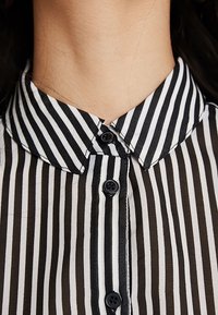 Stribet skjorte med knapper, med sorte og hvide lodrette striber, en spids krave og to sorte knapper på frontpladen.